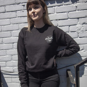Woman standing against a white painted brick wall wearing a fleece crew neck sweater with a screen printed, typography based graphic on the left chest