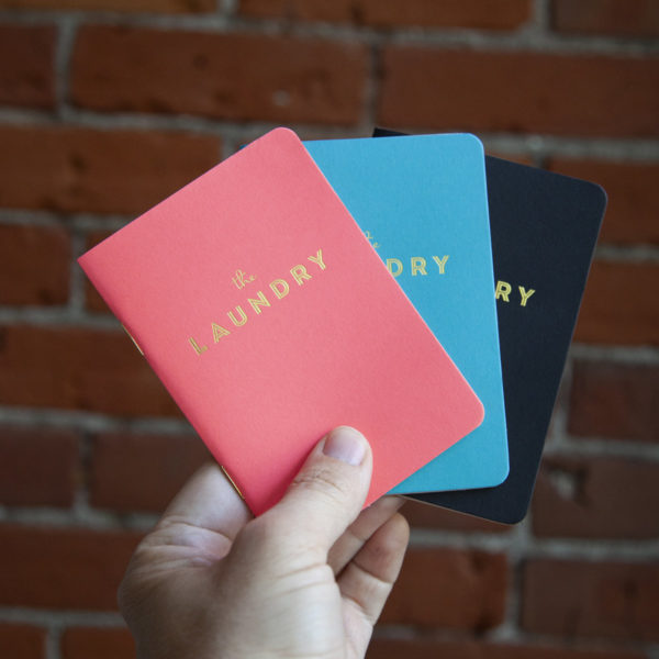 3 Pocket notebooks held up in a persons hand, coloured pink, blue, and black with gold text that reads "The Laundry"