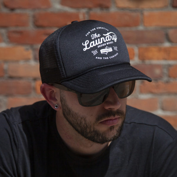 Man wearing sunglasses and a black retro foam trucker hat featuring a retro style Laundry Design Works logo graphic